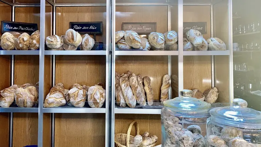 Étagères de pain : pain aux pois chiches, aux noix, aux figues, aux olives, baguettes, sur étagères en bois clair.