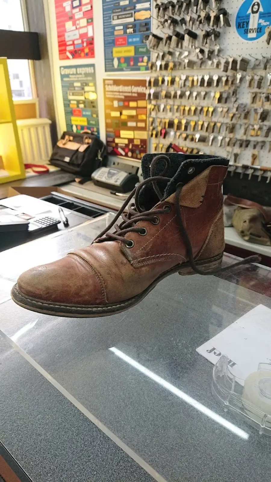 Botte en cuir brun en cours de préparation sur le comptoir, avec un mur d'étagères jaunes et bleues en arrière-plan rempli de clés et de panneaux KEY SERVICE et Schilderdienst.