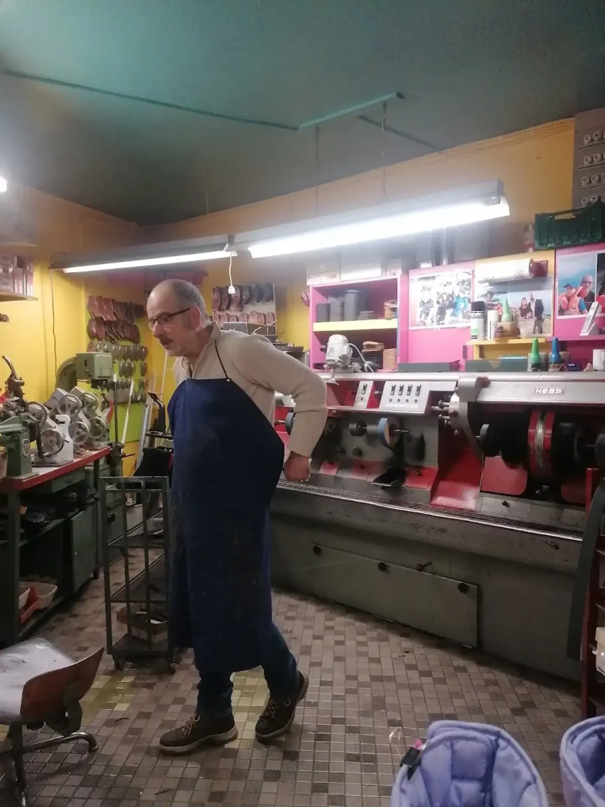 Angelo Veneziani au travail dans son atelier de cordonnerie : tablier bleu denim, machine professionnelle rouge en cours d'utilisation, plafond turquoise, murs jaunes vifs, étagères roses avec photos de famille.